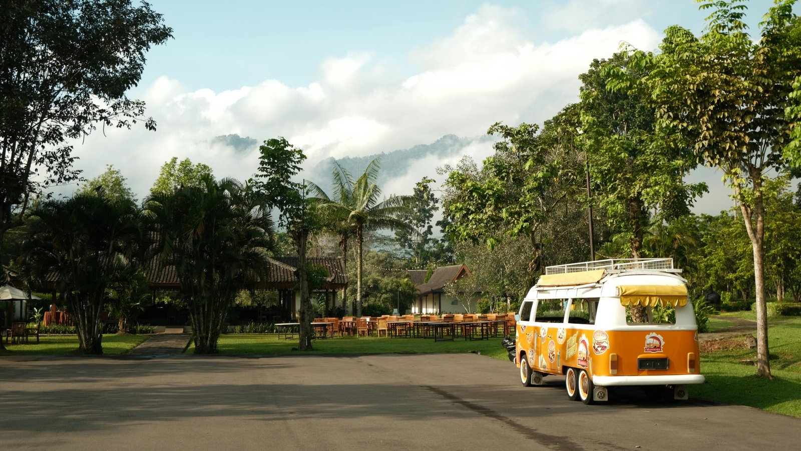 A vibrant orange camper van parked in a lush, tropical park under a blue sky.